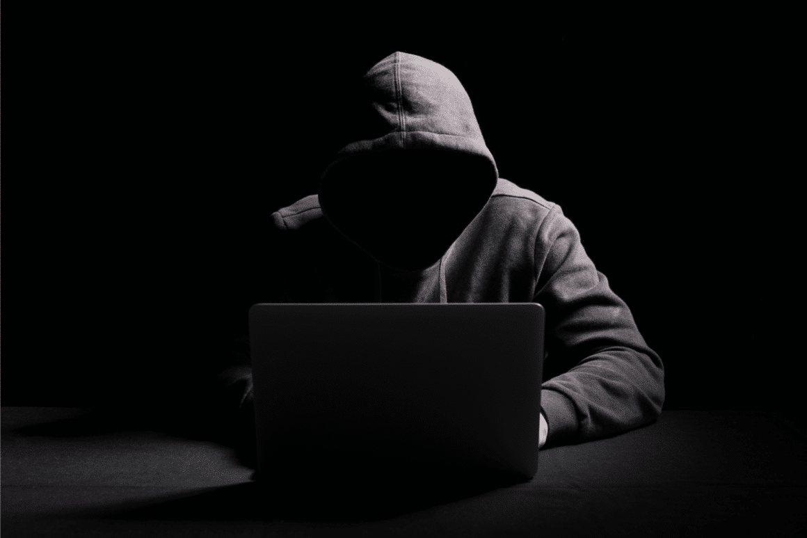 un hacker à capuche sans visage avec un laptop devant un fond noir