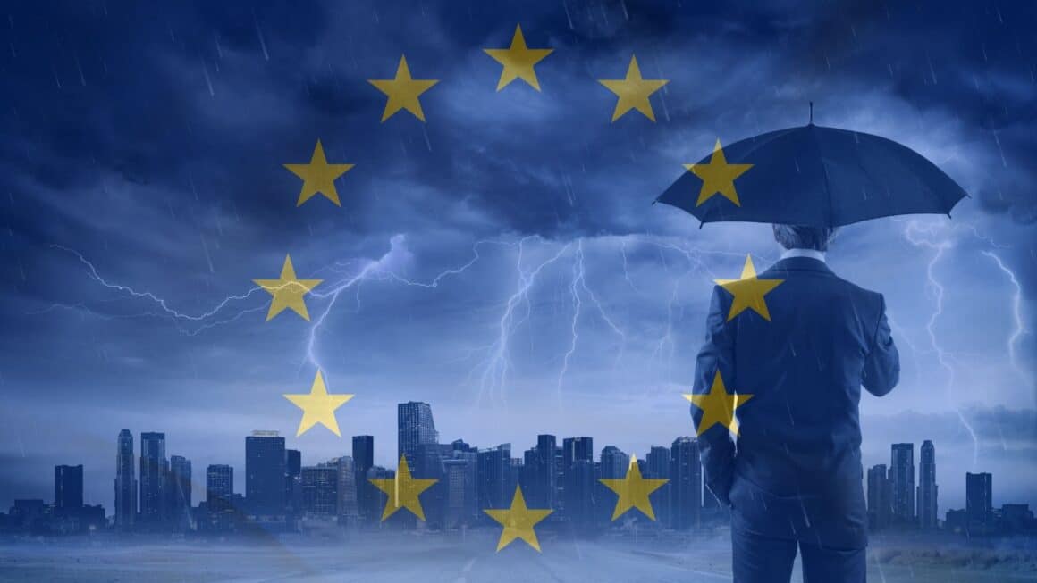 Un homme en costume vu de dos tenant un parapluie, face à une ville sous un orage violent. En surimpression, les étoiles du drapeau européen symbolisent la protection et la coordination de l’Union européenne face aux crises. L’ambiance visuelle évoque la cybersécurité et la résilience en contexte de menace.