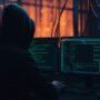 Personne de dos portant une capuche, assise devant plusieurs écrans affichant des lignes de code dans une salle sombre, illustrant une cyberattaque ou une activité de piratage informatique dans un environnement de cybersécurité.