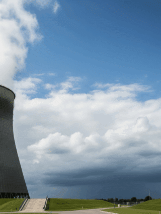 Tour de refroidissement d'une centrale nucléaire s'élevant vers un ciel bleu clair, avec des nuages sombres et menaçants à l'horizon, évoquant la puissance et la complexité de l'énergie nucléaire face aux défis environnementaux ou de sécurité.