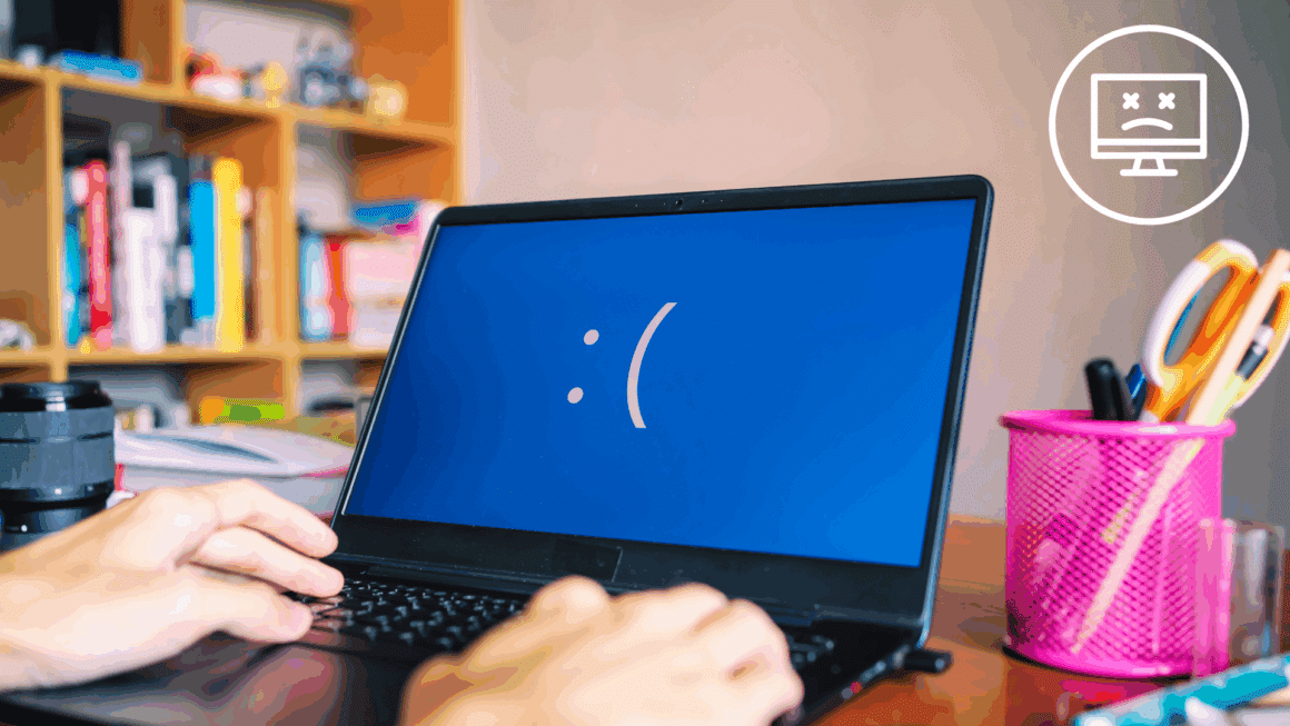 Gros plan d’un ordinateur portable affichant un écran bleu avec un visage triste, indiquant une erreur système critique, tandis qu'une personne tape au clavier dans un bureau à domicile avec des fournitures et des livres en arrière-plan.