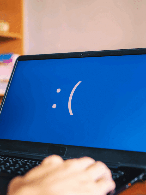 Gros plan d’un ordinateur portable affichant un écran bleu avec un visage triste, indiquant une erreur système critique, tandis qu'une personne tape au clavier dans un bureau à domicile avec des fournitures et des livres en arrière-plan.