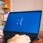 Gros plan d’un ordinateur portable affichant un écran bleu avec un visage triste, indiquant une erreur système critique, tandis qu'une personne tape au clavier dans un bureau à domicile avec des fournitures et des livres en arrière-plan.