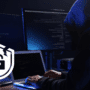 Personne portant un sweat à capuche devant plusieurs écrans affichant du code, représentant une cyberattaque en cours, avec une icône de sécurité cassée en surimpression.