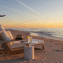 Une chaise longue luxueuse sur une plage de sable fin au coucher du soleil, face à une mer calme. La lumière chaude et dorée crée une ambiance sereine. Un avion de ligne vole dans le ciel en arrière-plan, évoquant le thème du voyage et des vacances.