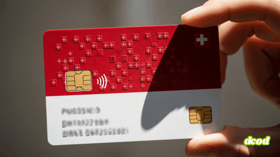 Gros plan sur une carte d'identité numérique aux couleurs de la Suisse (rouge et blanche), tenue entre deux doigts. La carte est dotée d'une puce électronique et du symbole de la croix suisse.