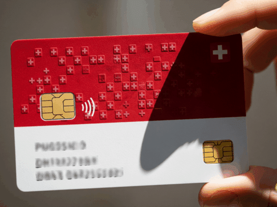 Gros plan sur une carte d'identité numérique aux couleurs de la Suisse (rouge et blanche), tenue entre deux doigts. La carte est dotée d'une puce électronique et du symbole de la croix suisse.