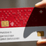 Gros plan sur une carte d'identité numérique aux couleurs de la Suisse (rouge et blanche), tenue entre deux doigts. La carte est dotée d'une puce électronique et du symbole de la croix suisse.