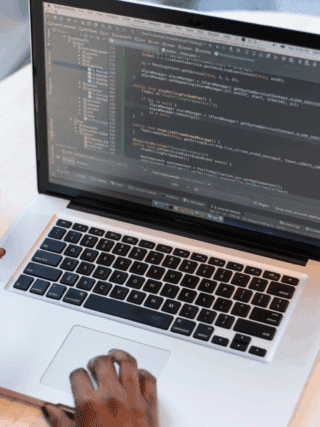 Vue rapprochée d’un développeur travaillant sur une application Android avec un ordinateur portable affichant du code Java dans Android Studio, tout en testant l’application sur un smartphone connecté via USB. Une tasse de café, une paire de lunettes et le logo Android sont visibles sur la table.