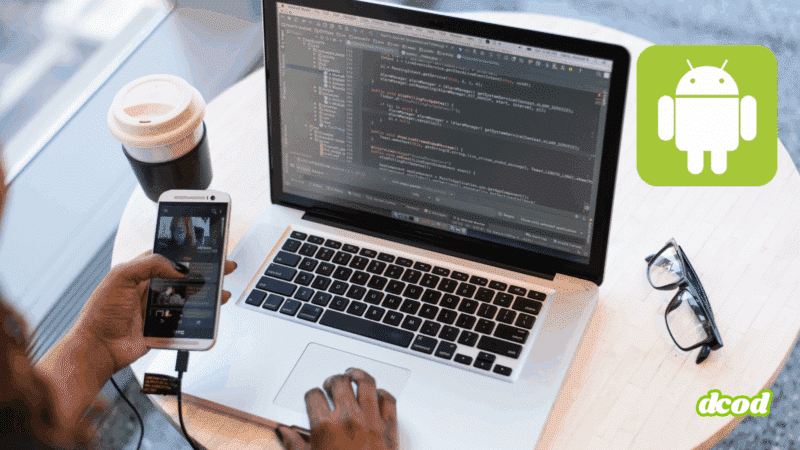 Vue rapprochée d’un développeur travaillant sur une application Android avec un ordinateur portable affichant du code Java dans Android Studio, tout en testant l’application sur un smartphone connecté via USB. Une tasse de café, une paire de lunettes et le logo Android sont visibles sur la table.