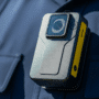 Gros plan hyper-réaliste d'une bodycam noire attachée à l'uniforme bleu d'un policier, mettant en évidence les détails de l'appareil.