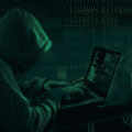 Personne portant une capuche, tapant sur un ordinateur portable dans un décor sombre, illustrant le piratage informatique ou une cyberattaque, avec des lignes de code visibles à l’écran.