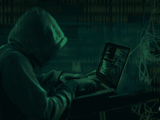 Personne portant une capuche, tapant sur un ordinateur portable dans un décor sombre, illustrant le piratage informatique ou une cyberattaque, avec des lignes de code visibles à l’écran.
