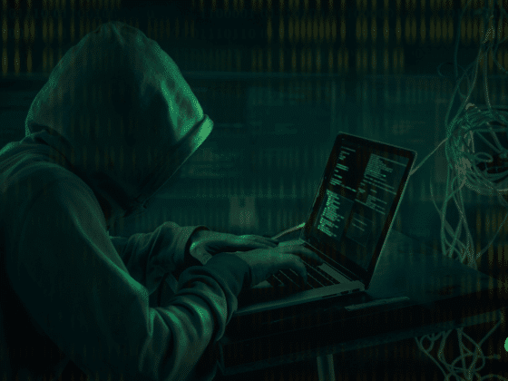 Personne portant une capuche, tapant sur un ordinateur portable dans un décor sombre, illustrant le piratage informatique ou une cyberattaque, avec des lignes de code visibles à l’écran.
