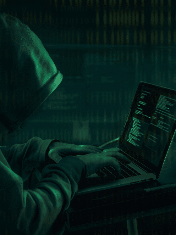 Personne portant une capuche, tapant sur un ordinateur portable dans un décor sombre, illustrant le piratage informatique ou une cyberattaque, avec des lignes de code visibles à l’écran.