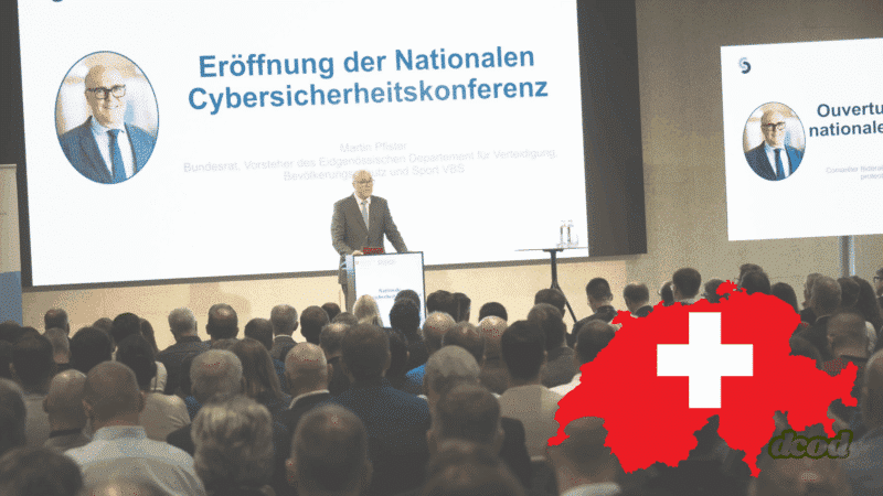 Martin Pfister, conseiller fédéral suisse, prononce le discours d’ouverture de la Conférence Nationale sur la Cybersécurité devant un public attentif, avec une carte rouge de la Suisse ornée d'une croix blanche en premier plan.
