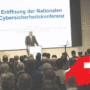 Martin Pfister, conseiller fédéral suisse, prononce le discours d’ouverture de la Conférence Nationale sur la Cybersécurité devant un public attentif, avec une carte rouge de la Suisse ornée d'une croix blanche en premier plan.