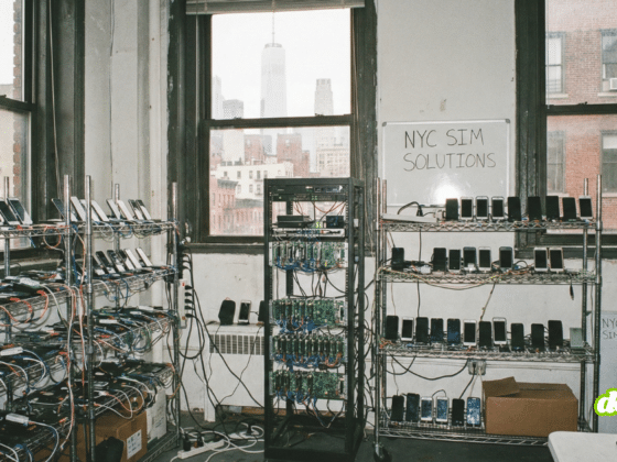 Photographie réaliste au format paysage montrant l'intérieur d'un local industriel new-yorkais reconverti en ferme à cartes SIM. Des rayonnages métalliques sont remplis de centaines de smartphones alignés et connectés par un enchevêtrement de câbles. L'espace est brut avec des murs usés, et de grandes fenêtres industrielles en arrière-plan révèlent une vue urbaine typique de New York avec des bâtiments en briques rouges.