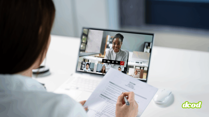 Vue arrière d’une personne tenant un CV en main lors d’un entretien d’embauche en visioconférence sur un ordinateur portable, avec six participants visibles à l’écran dans un environnement de bureau lumineux.