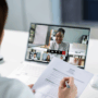 Vue arrière d’une personne tenant un CV en main lors d’un entretien d’embauche en visioconférence sur un ordinateur portable, avec six participants visibles à l’écran dans un environnement de bureau lumineux.
