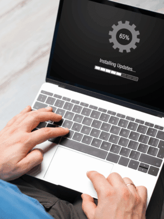 Vue en plongée d’un homme utilisant un ordinateur portable affichant une mise à jour en cours (65 %), illustrant un processus de mise à jour système conforme aux recommandations de cybersécurité du NIST, avec les logos NIST et dcod visibles à l’écran