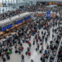 Vue en plongée d'un vaste hall d'aéroport lumineux et bondé, où une foule de voyageurs avec leurs bagages circule entre les comptoirs d'enregistrement et les escalators, sous une structure architecturale moderne.