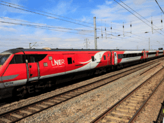 Train à grande vitesse rouge et gris de la compagnie LNER (London North Eastern Railway) circulant sur une voie ferrée électrifiée par une journée ensoleillée, au milieu d’un paysage rural britannique.