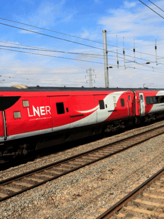 Train à grande vitesse rouge et gris de la compagnie LNER (London North Eastern Railway) circulant sur une voie ferrée électrifiée par une journée ensoleillée, au milieu d’un paysage rural britannique.
