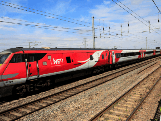 Train à grande vitesse rouge et gris de la compagnie LNER (London North Eastern Railway) circulant sur une voie ferrée électrifiée par une journée ensoleillée, au milieu d’un paysage rural britannique.