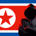 Personne en sweat à capuche noire, visage masqué par l’ombre, symbolisant un hacker, positionnée devant le drapeau de la Corée du Nord, représentant la cybercriminalité ou les activités de piratage informatique attribuées au pays.