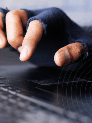 Gros plan sur une main portant un gant sans doigts tapant sur un clavier d’ordinateur, symbolisant une activité potentiellement illégale ou anonyme liée au Bitcoin, représenté par son logo orange en bas à gauche. Logo « dcod » en bas à droite.