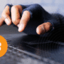 Gros plan sur une main portant un gant sans doigts tapant sur un clavier d’ordinateur, symbolisant une activité potentiellement illégale ou anonyme liée au Bitcoin, représenté par son logo orange en bas à gauche. Logo « dcod » en bas à droite.