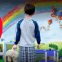 Jeune garçon en pyjama tenant une peluche, debout face à une fresque colorée représentant un arc-en-ciel et des éléments joyeux d’enfance, avec en surimpression un personnage de hacker en capuche sur fond rouge, symbolisant les risques de cybercriminalité ciblant les enfants.