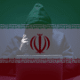 Hacker anonyme en sweat à capuche, assis devant un ordinateur portable, sur fond du drapeau iranien semi-transparent, symbolisant une cyberattaque ou une menace informatique liée à l’Iran.