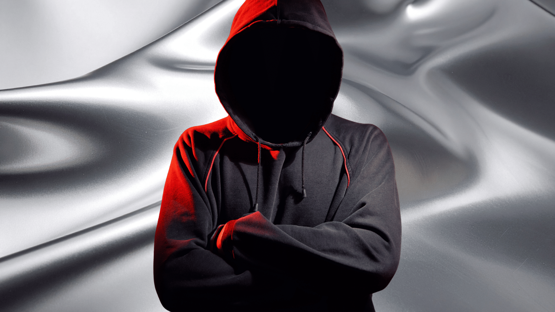 Personne non identifiable portant un sweat à capuche noir et rouge, bras croisés, avec le visage dissimulé dans l’ombre, posant devant un fond métallique fluide, symbolisant l’anonymat et la menace cyber dans un univers numérique.