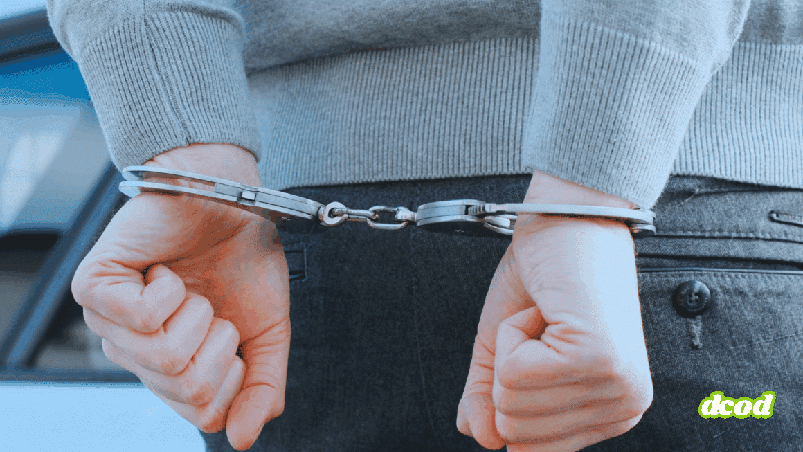 Gros plan sur les mains d'une personne menottée dans le dos, en jean et pull gris, illustrant une arrestation par les forces de l'ordre.