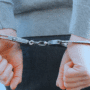 Gros plan sur les mains d'une personne menottée dans le dos, en jean et pull gris, illustrant une arrestation par les forces de l'ordre.