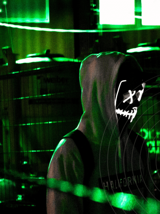 Personne portant un sweat à capuche et un masque lumineux stylisé de type "hacker", se tenant dans un environnement urbain sombre éclairé par des néons verts, avec des grilles métalliques en arrière-plan et le logo « dcod » en bas à droite de l’image.