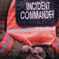 Vue rapprochée d’un commandant d’incident en tenue de gestion de crise, les mains croisées dans le dos, avec un gilet haute visibilité marqué « Incident Commander », sur fond de code informatique symbolisant la cybersécurité.