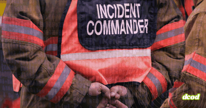 Vue rapprochée d’un commandant d’incident en tenue de gestion de crise, les mains croisées dans le dos, avec un gilet haute visibilité marqué « Incident Commander », sur fond de code informatique symbolisant la cybersécurité.