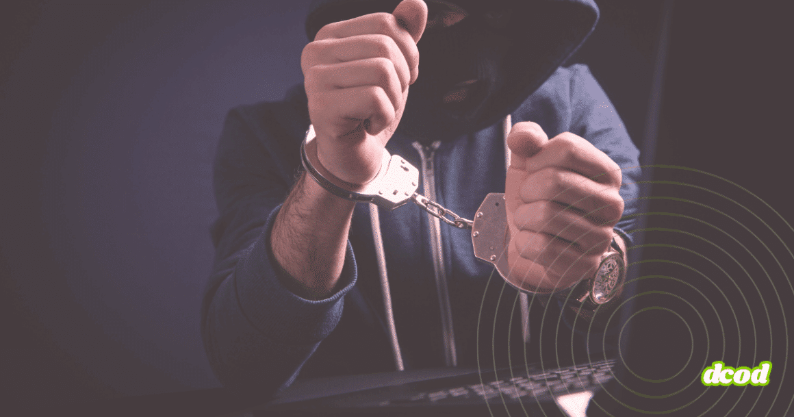 Personne cagoulée en sweat à capuche, menottée devant un clavier d’ordinateur, illustrant l’arrestation d’un cybercriminel.