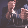 Personne cagoulée en sweat à capuche, menottée devant un clavier d’ordinateur, illustrant l’arrestation d’un cybercriminel.