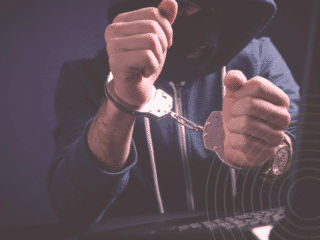 Personne cagoulée en sweat à capuche, menottée devant un clavier d’ordinateur, illustrant l’arrestation d’un cybercriminel.
