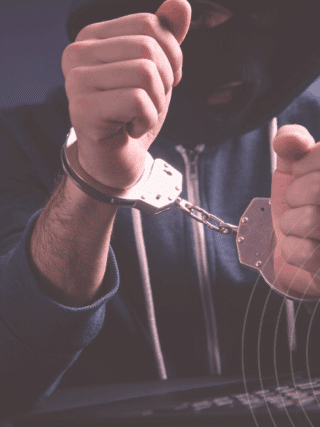 Personne cagoulée en sweat à capuche, menottée devant un clavier d’ordinateur, illustrant l’arrestation d’un cybercriminel.
