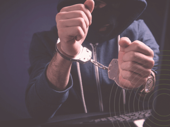 Personne cagoulée en sweat à capuche, menottée devant un clavier d’ordinateur, illustrant l’arrestation d’un cybercriminel.