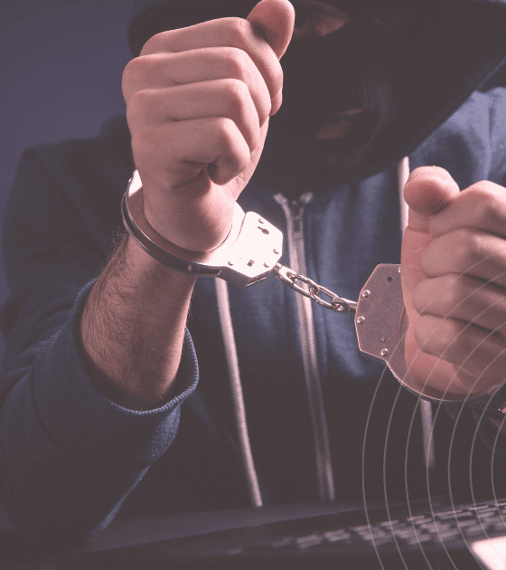 Personne cagoulée en sweat à capuche, menottée devant un clavier d’ordinateur, illustrant l’arrestation d’un cybercriminel.