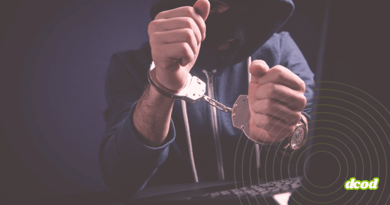 Personne cagoulée en sweat à capuche, menottée devant un clavier d’ordinateur, illustrant l’arrestation d’un cybercriminel.