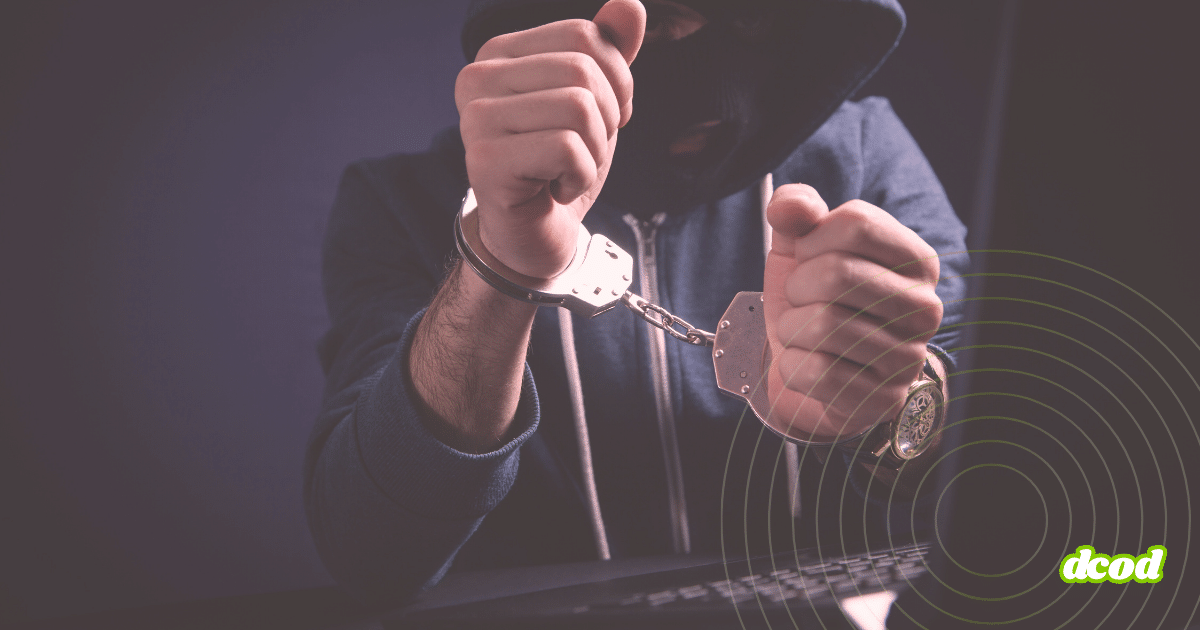 Personne cagoulée en sweat à capuche, menottée devant un clavier d’ordinateur, illustrant l’arrestation d’un cybercriminel.