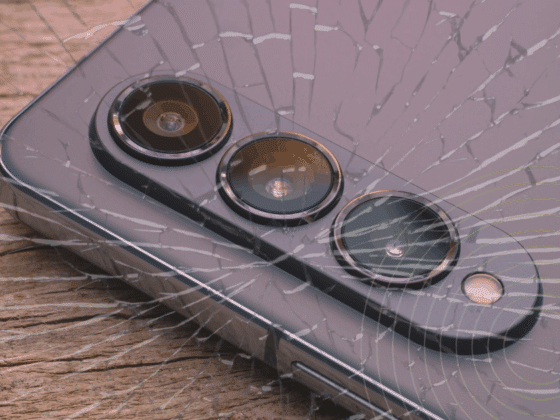 Gros plan sur un smartphone moderne avec triple capteur photo, posé sur du bois, recouvert d’un effet de verre fissuré symbolisant une faille ou une vulnérabilité. Logo "dcod" en bas à droite.
