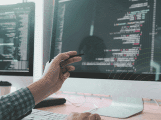 Développeur travaillant sur un ordinateur double écran affichant du code source, avec une main tenant un stylet pointant vers l'écran principal. Atmosphère de bureau technique et logo "dcod" visible en bas à droite.